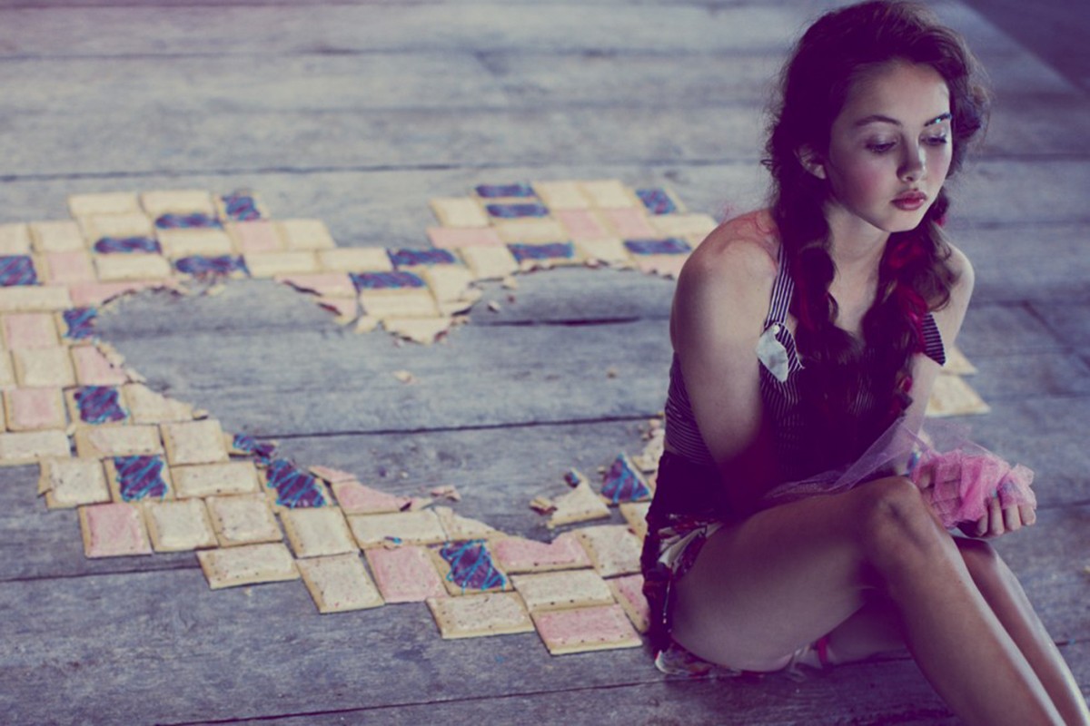 A young woman with braids sits near a heart made of crackers on a wooden floor in soft, diffused light, creating a whimsical atmosphere.