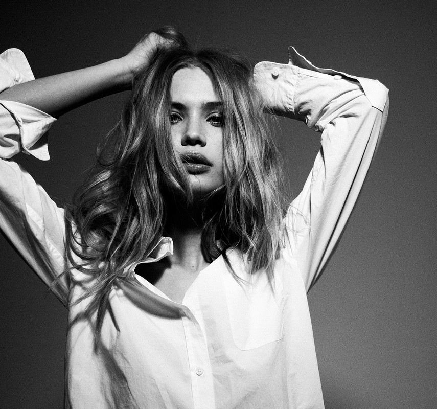 Monochrome portrait of a woman with tousled hair wearing a white button-down shirt with her hands up by her head against a dark grey background.