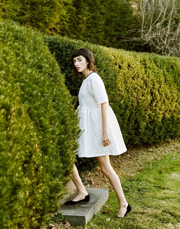 A woman in a white dress and black shoes walks on concrete steps amidst green hedges in a daytime outdoor fashion shoot.