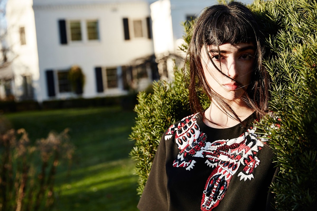Close-up of an adult female with dark hair standing near a green bush, wearing a black top with decorative red and white accents.