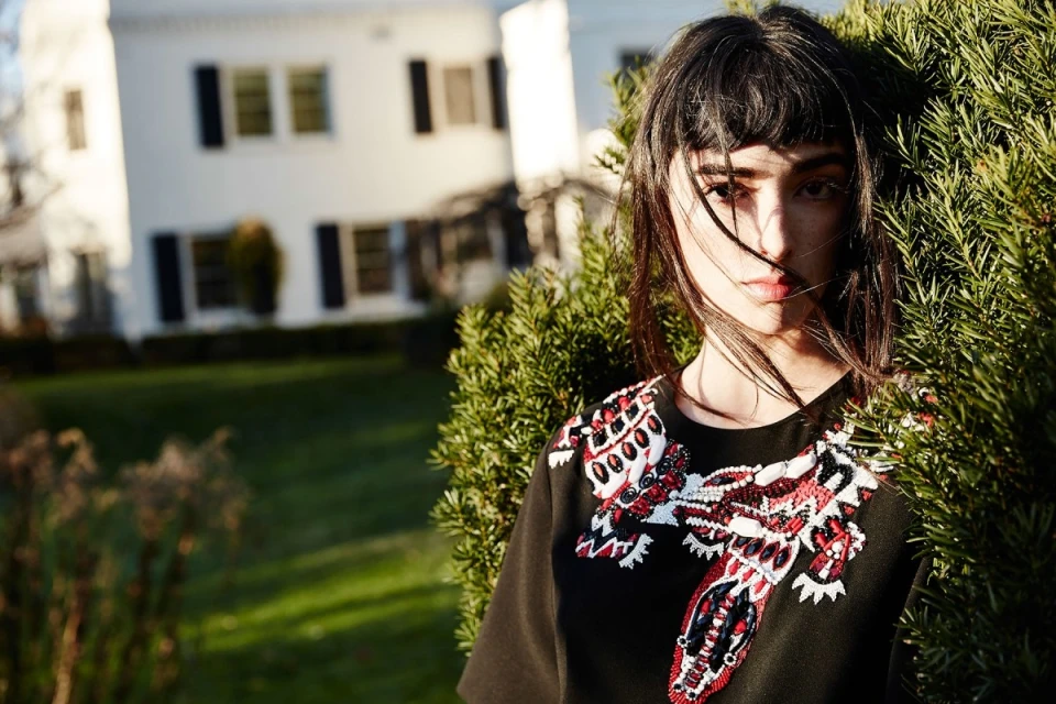 Close-up of an adult female with dark hair standing near a green bush, wearing a black top with decorative red and white accents.