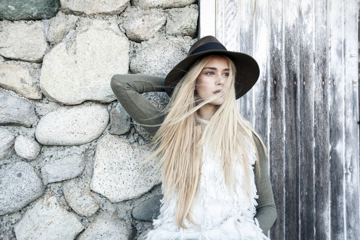 A blonde woman wearing a hat and a fringed top poses against a stone wall and a weathered wooden door in an outdoor setting.