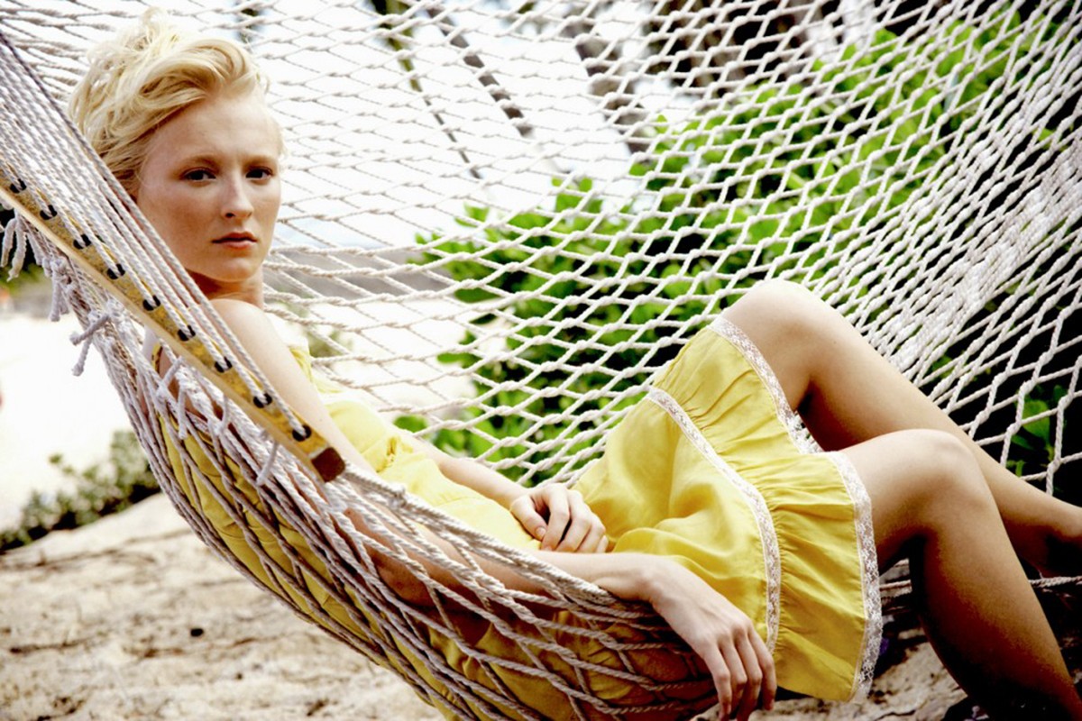 Woman relaxing in a hammock outdoors, wearing a yellow dress. The hammock is white, with a blurred background of trees and sand.