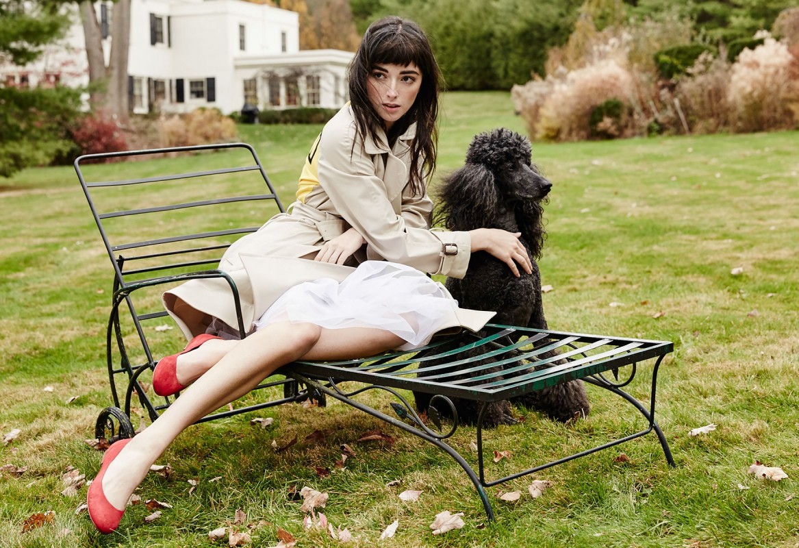 Woman in trench coat reclines on metal chair with black poodle in a grassy yard with a white house and trees in the background.