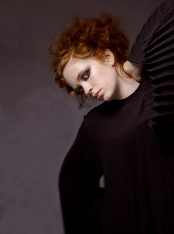 Portrait of a red-haired woman with dark eye makeup wearing a black top, posing against a muted gray background in studio lighting.
