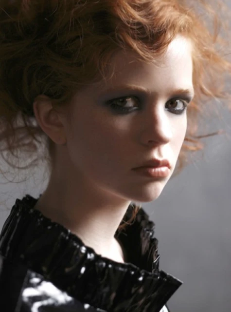 Close-up portrait of a red-haired woman with smoky eye makeup wearing a high-gloss black ruffled collar against a neutral grey background.
