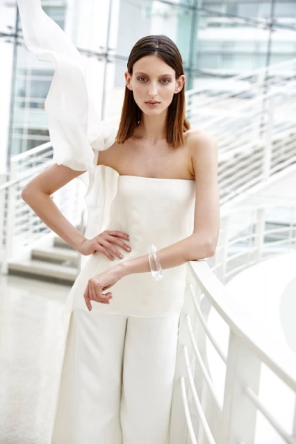 Fashion shot of a slender woman in white palazzo pants and strapless top with arm resting on a white railing, looking at camera.
