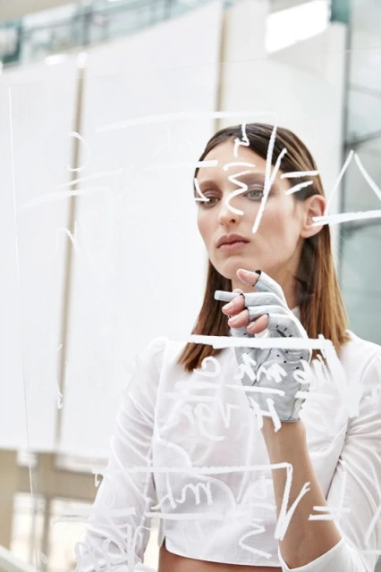 Woman in white shirt writing on glass wall, wearing gray prosthetic hand, indoor setting with bright lighting, medium shot.