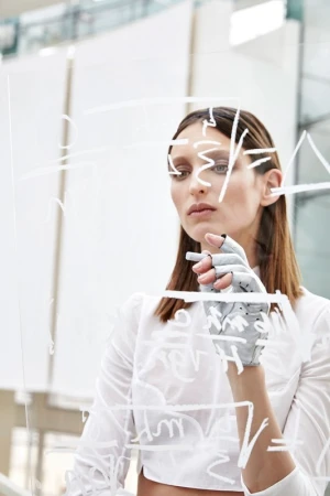 Woman in white shirt writing on glass wall, wearing gray prosthetic hand, indoor setting with bright lighting, medium shot.
