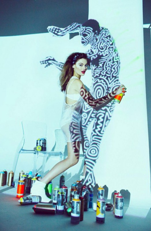 Adult woman poses with black and white patterned projected image and spray paint, illuminated by blue and white lighting in a photo studio.