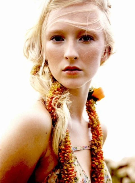 Portrait of a fair-skinned woman with blonde hair wearing a floral dress and flower garland, set against a bright outdoor background.