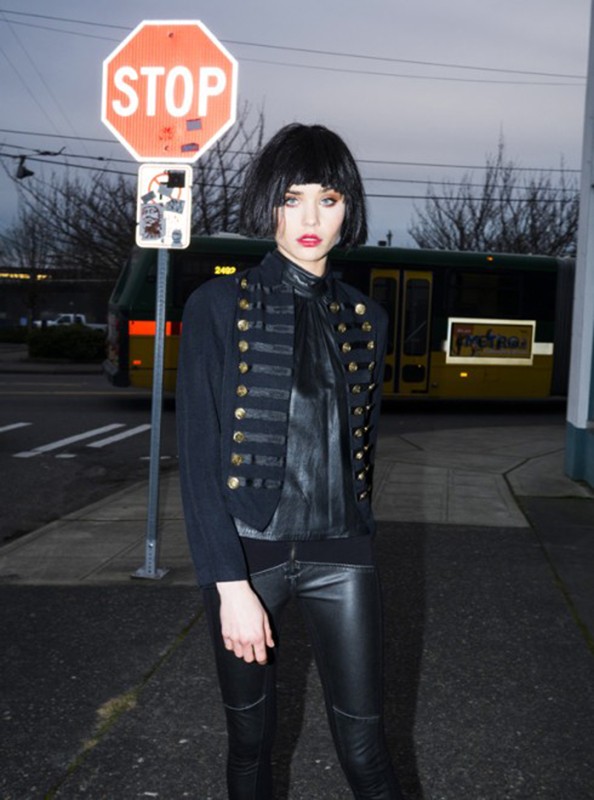 Adult woman poses in black jacket and leather pants next to a stop sign on city street with a bus in the background at dusk.