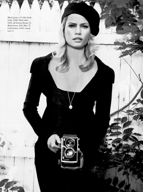 Black and white shot of a blonde woman in a beret and black suit, holding a vintage camera in an outdoor, leafy setting.