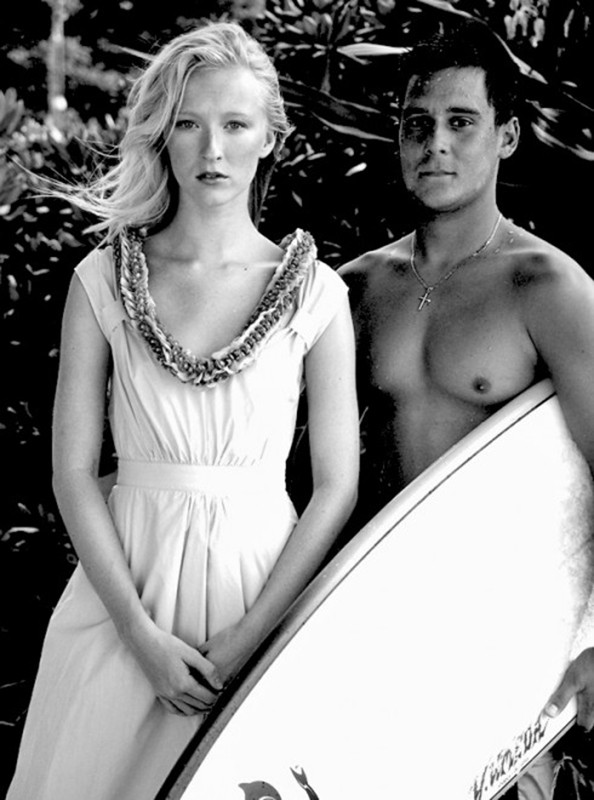 Black and white image of a woman in a light dress with a floral collar and a shirtless man holding a surfboard outdoors.