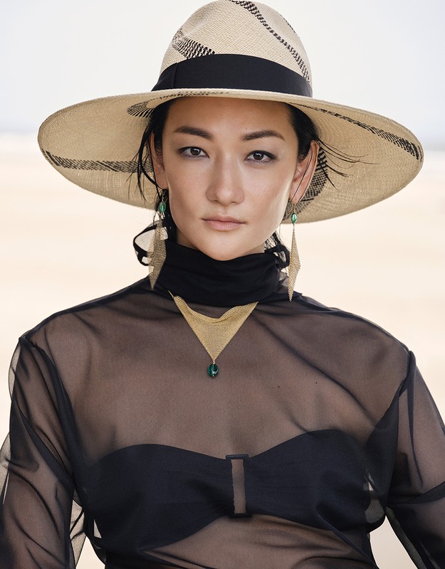 Portrait of an Asian woman wearing a wide-brimmed hat, sheer top, and geometric jewelry against a neutral sandy background.