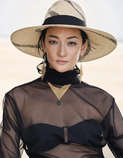 Portrait of an Asian woman wearing a wide-brimmed hat, sheer top, and geometric jewelry against a neutral sandy background.