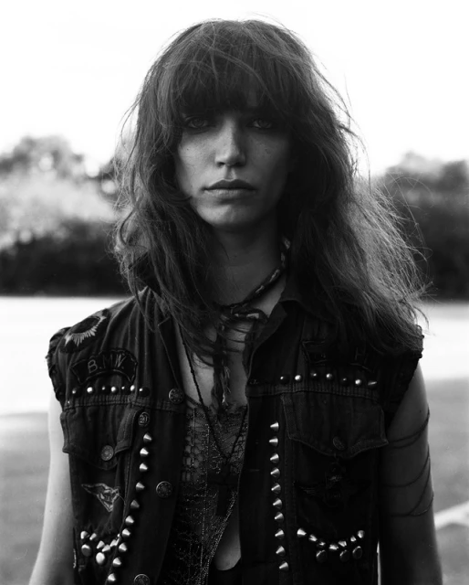 Black and white portrait of a woman with long, wavy hair wearing a studded denim vest in an outdoor setting.