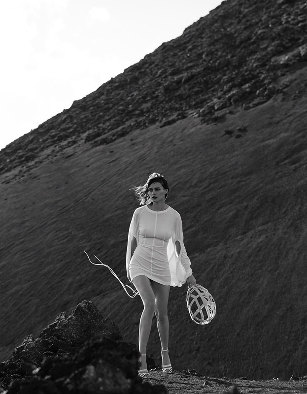 Woman in white dress poses on rocky terrain near large hill, holding decorative orb and sticks, in black and white outdoor fashion shot.