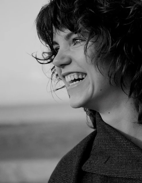 Smiling woman in a dark patterned jacket, side profile, set against a blurred gray outdoor background in a black and white portrait.