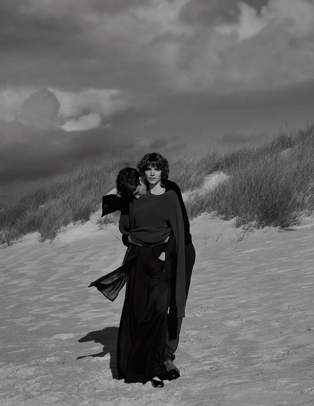 Monochrome image of a woman being carried by a man on a sandy dune with cloudy skies above, creating a dramatic, romantic visual narrative.