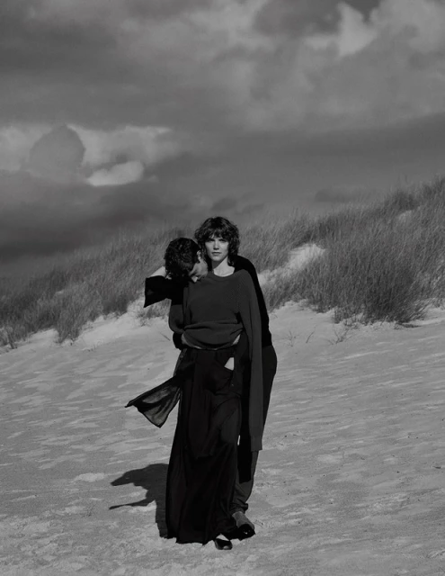 Monochrome image of a woman being carried by a man on a sandy dune with cloudy skies above, creating a dramatic, romantic visual narrative.