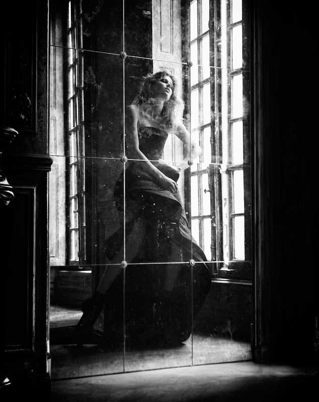 Elegant woman in strapless gown gazes out window reflected in paneled mirror in darkened interior, high contrast black and white image.