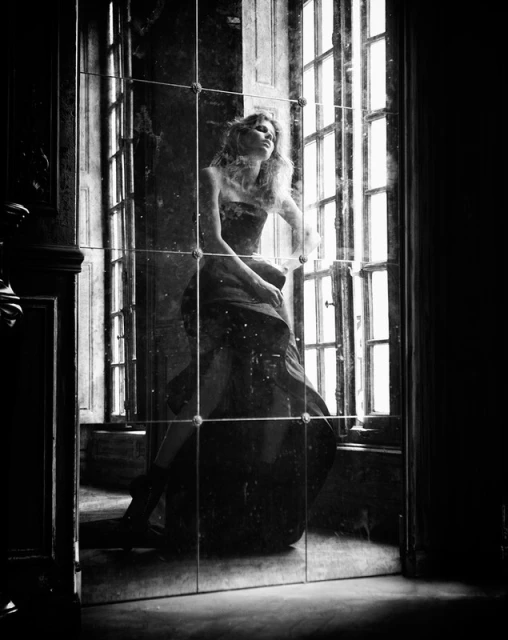 Elegant woman in strapless gown gazes out window reflected in paneled mirror in darkened interior, high contrast black and white image.