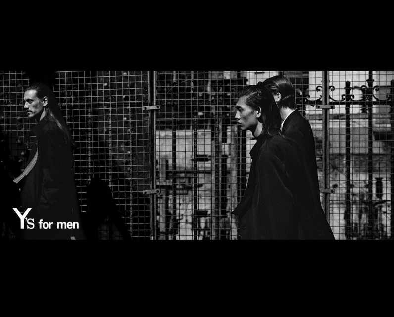 A black and white fashion photograph showcases three men in dark suits walking past a metal grate in an outdoor urban environment.
