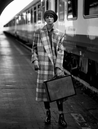 Woman in plaid coat with vintage suitcase stands on train platform in a black and white editorial fashion photograph.