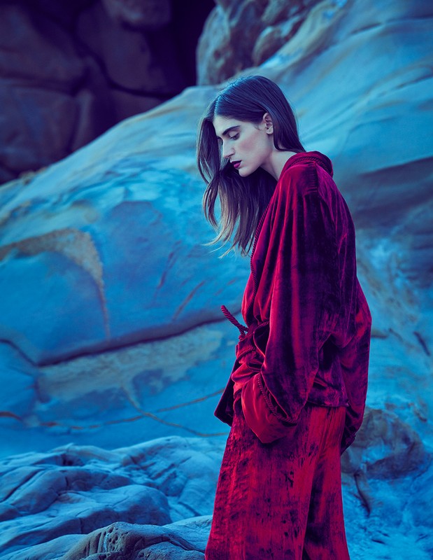A woman in a magenta outfit stands in profile against a blue rock formation, looking downward in an outdoor setting with soft lighting.