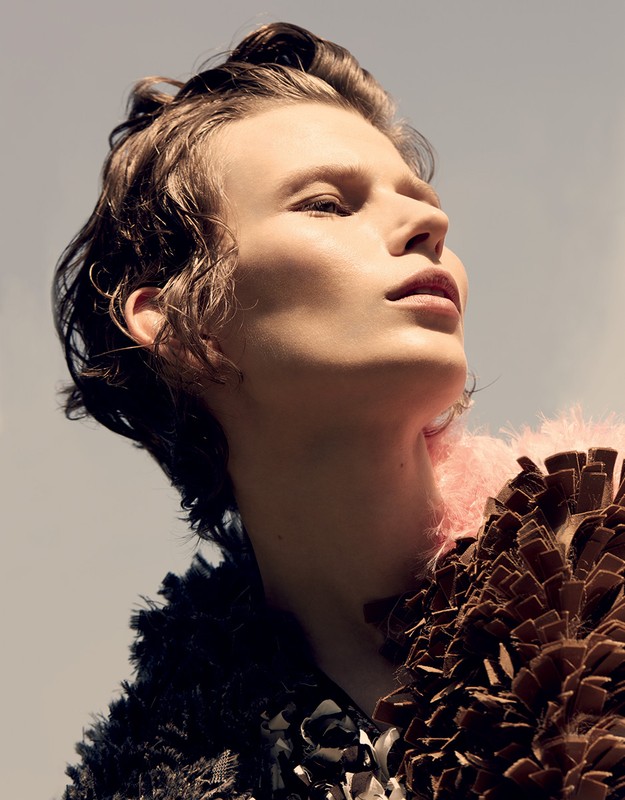 Fashion shot of a fair-skinned adult looking up, featuring a structured brown and black garment with a pink feathered collar under blue sky.
