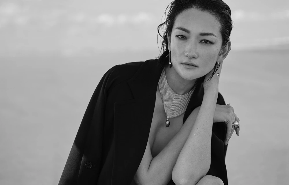 Striking black and white fashion portrait of an Asian woman with wet hair wearing pearl jewelry and a black blazer against a light sky.