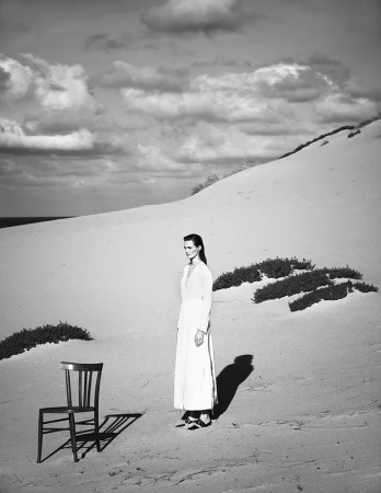Black and white editorial fashion photograph of a woman standing on a sand dune next to a wooden chair under a cloudy sky.