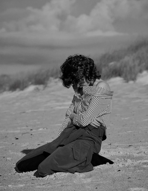 Monochrome portrait of a woman with short curly hair, kneeling on a beach with dunes and cloudy sky behind.