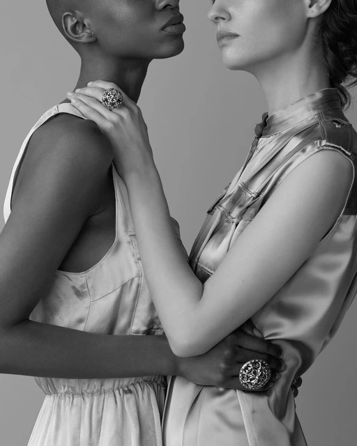 Two women of different ethnicities pose together in a studio, showcasing large ornate rings. Black and white, high-key lighting.