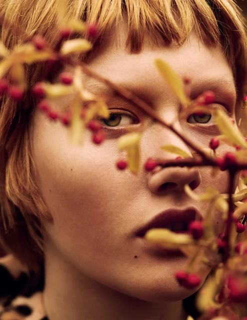 Close-up portrait of a young adult woman with red-blonde hair, partially obscured by branches with red berries and yellow leaves.
