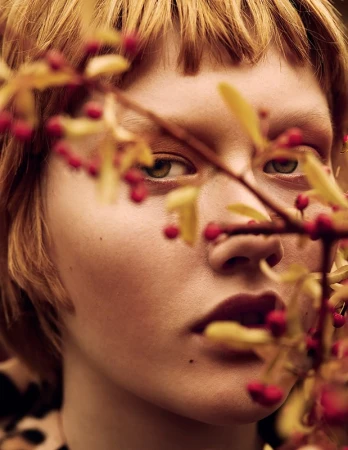 Close-up portrait of a young adult woman with red-blonde hair, partially obscured by branches with red berries and yellow leaves.
