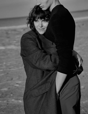 Black-and-white portrait of a hugging couple on a beach, woman with short brown hair in tweed jacket and man in black sweater.