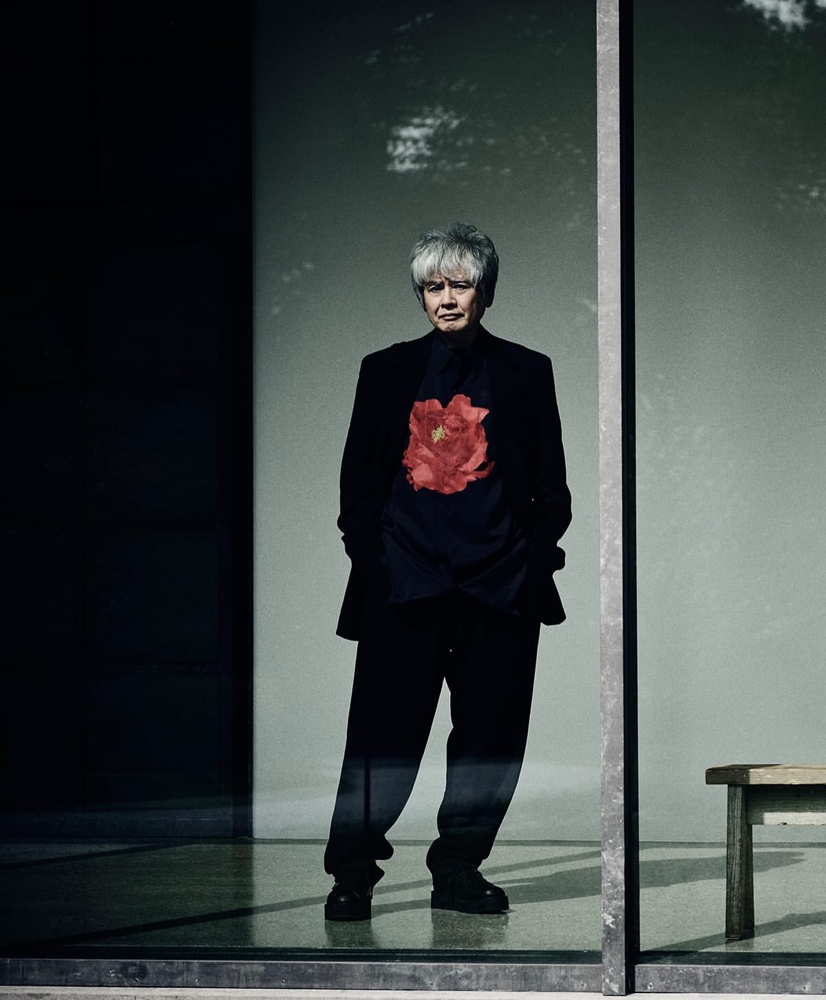 Older adult stands in front of a window wearing a black floral shirt, black pants, and black blazer inside an architectural structure.