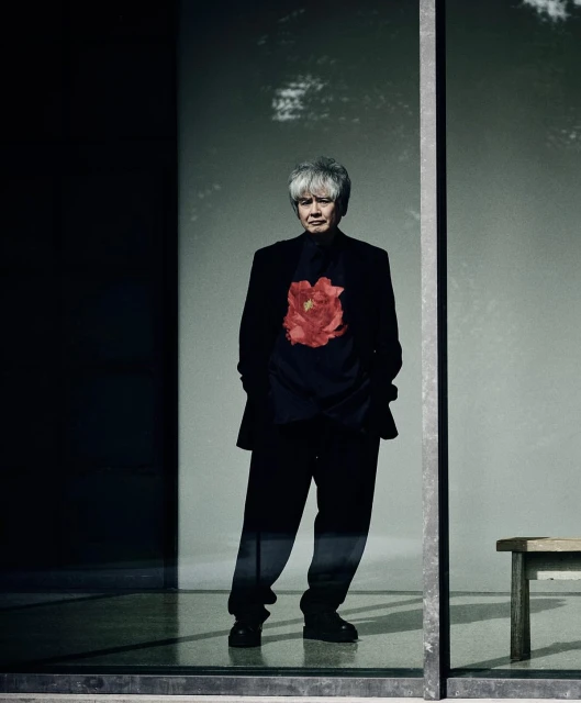 Older adult stands in front of a window wearing a black floral shirt, black pants, and black blazer inside an architectural structure.