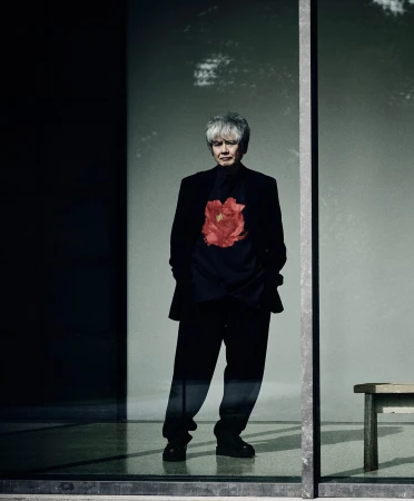 Older adult stands in front of a window wearing a black floral shirt, black pants, and black blazer inside an architectural structure.