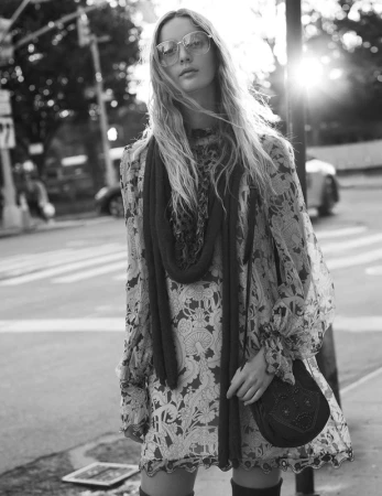 Monochrome street portrait of a woman wearing patterned dress, oversized glasses, scarf, and carrying shoulder bag
