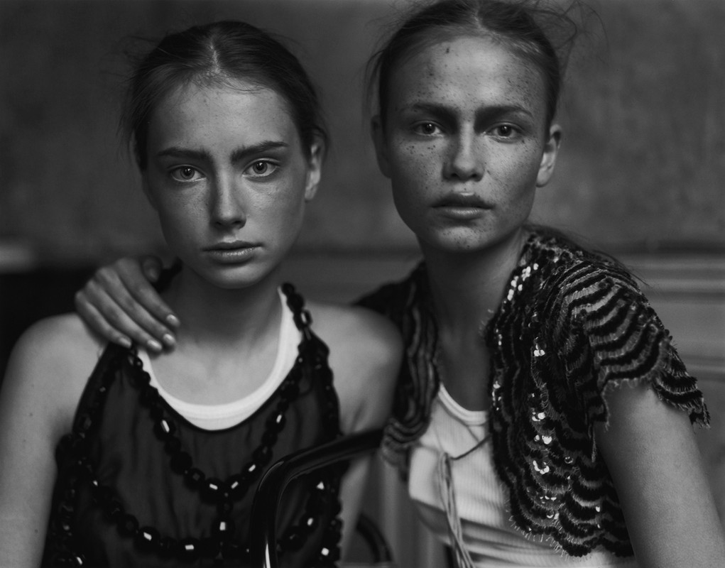 Monochromatic portrait of two fair-skinned young women with light brown hair, wearing casual tops and looking directly at the camera.