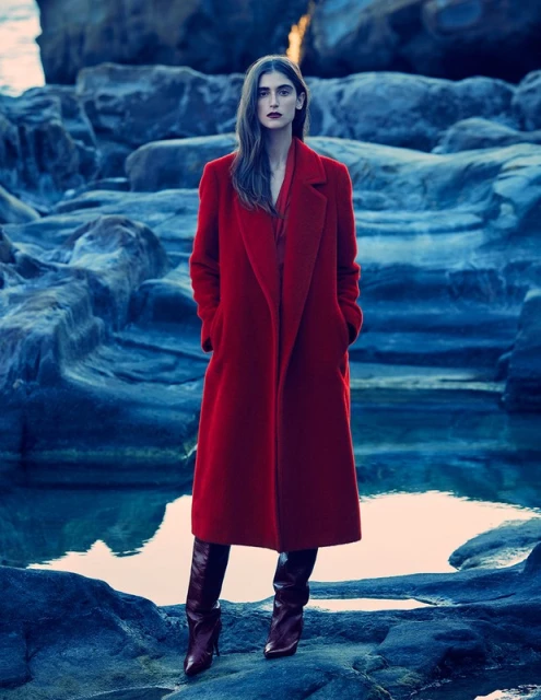 Elegant woman in a long red coat and boots posing outdoors against a rocky coastal backdrop and standing near the water's edge.