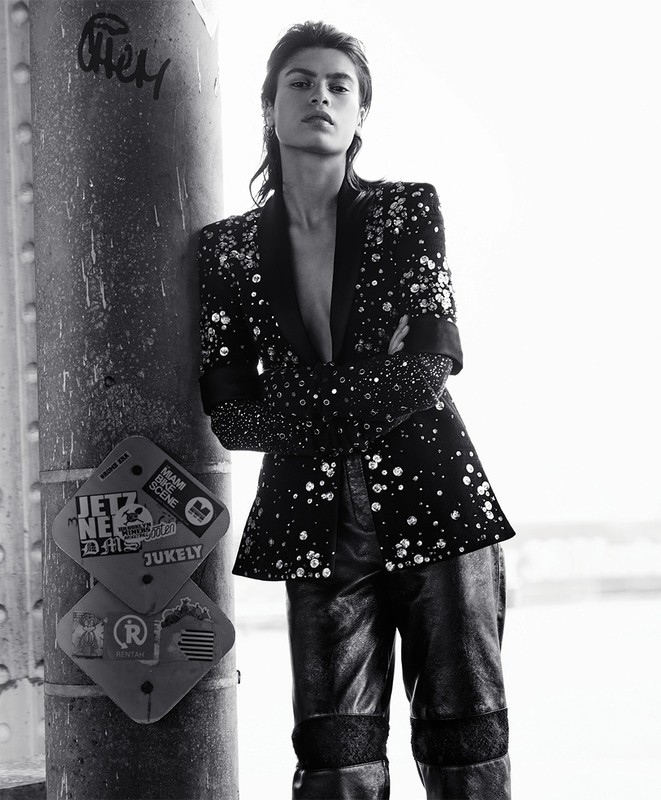 Monochrome fashion portrait of a stylish woman leaning against a pillar wearing a blazer and pants with embellishments looking at the camera.