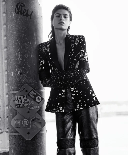 Monochrome fashion portrait of a stylish woman leaning against a pillar wearing a blazer and pants with embellishments looking at the camera.