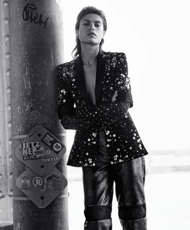 Monochrome fashion portrait of a stylish woman leaning against a pillar wearing a blazer and pants with embellishments looking at the camera.