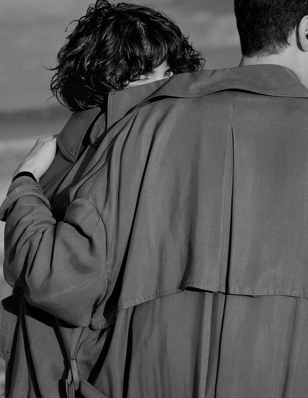 Black and white portrait of a woman partially obscured by a trench coat, standing beside a man against an outdoor background.