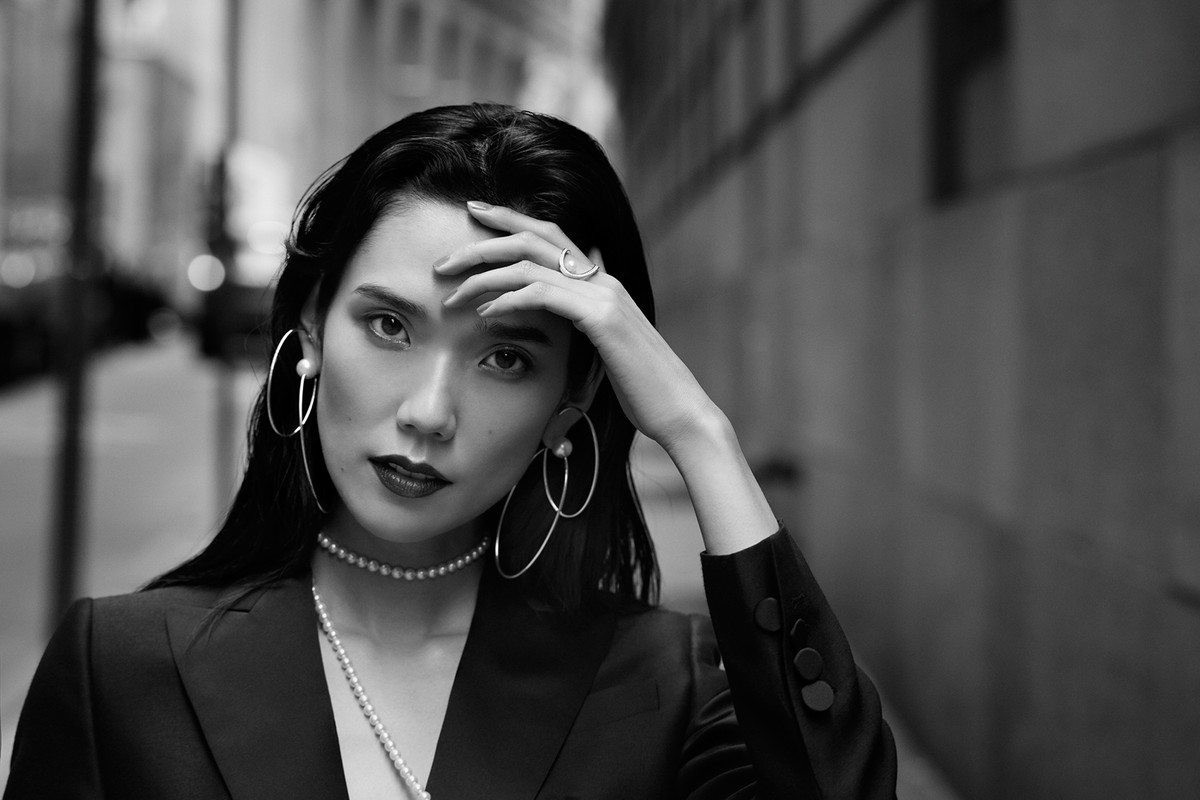 Black and white portrait of an Asian woman with large hoop earrings, pearl necklace, and a blazer against an urban background.