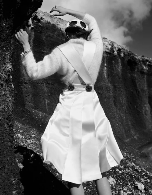 Black and white fashion shot from the back of an adult woman wearing a white sweater and skirt while standing outdoors on a rock.